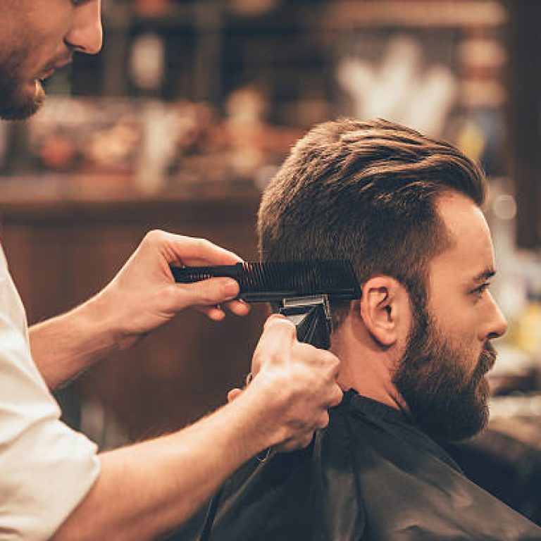 Close up side view of young bearded man getting haircut by hairdresser with electric razor at barbershop
